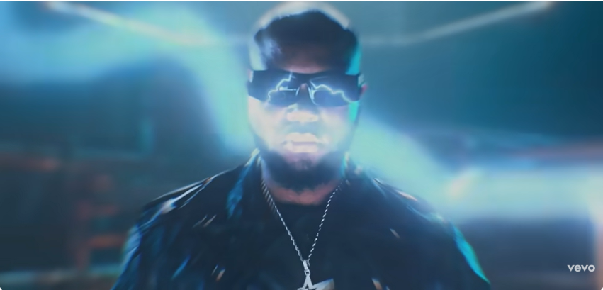 Screenshot: King Promise as the 'Terminator' in sci-fi mode King Promise in dramatic blue lighting, wearing futuristic black sunglasses with white-blue lightning arcing across the lenses, a dark jacket, and silver star pendant.