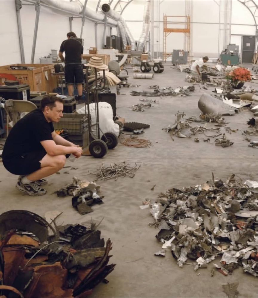 Elon Musk staring at a bunch of scrap metal while squatting